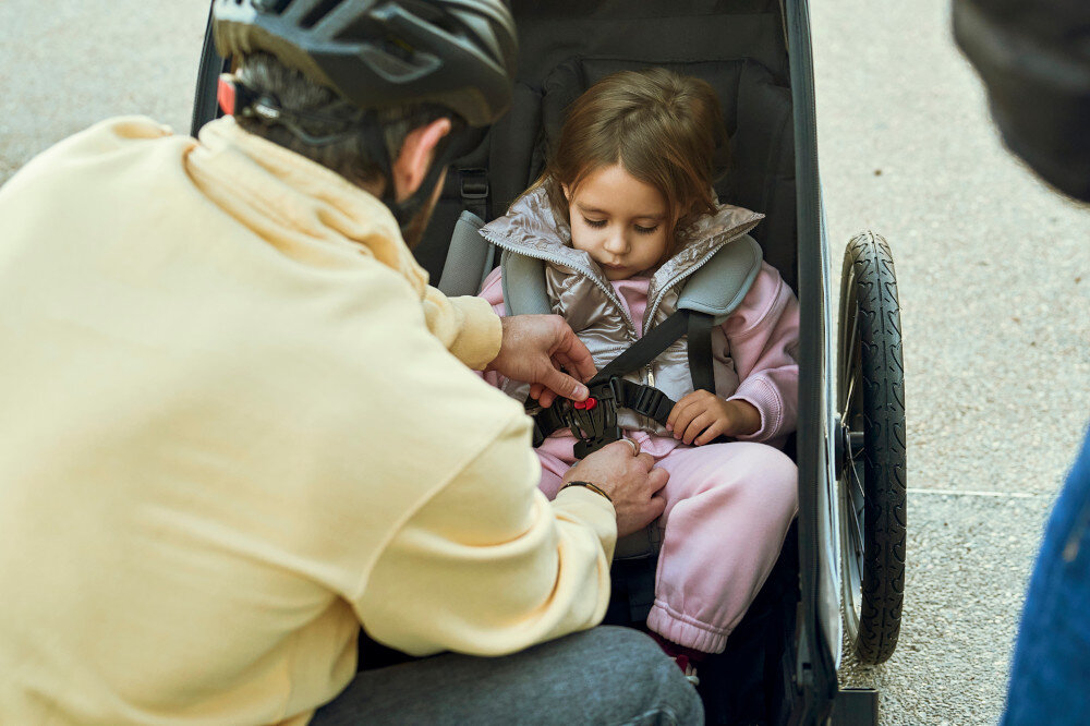 Przyczepka rowerowa KINDERKRAFT Nava Walk 2w1 60kg Pasy Czarno-szary przyczepka na białym tle widok na wnętrze 5-punktowe pasy bezpieczeństwa solidna podłoga aluminiowa rama przyciemniane boki osłona przeciwdeszczowa przeciwsłoneczna moskitiera odblaski flaga ostrzegawcza dwa hamulce pełna kontrola nad wózkiem