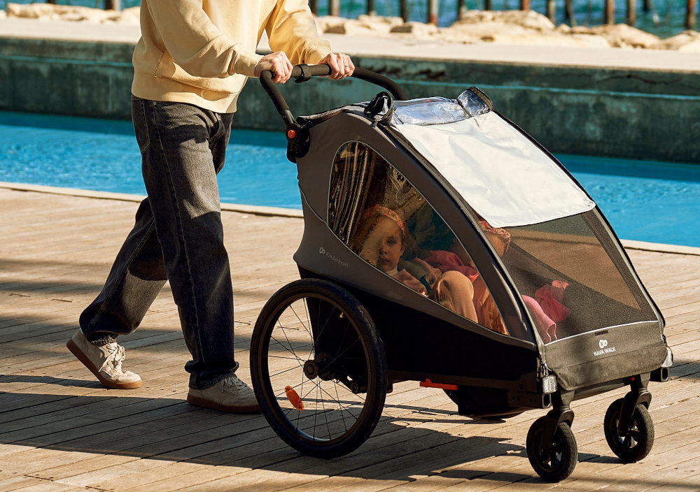 Przyczepka rowerowa KINDERKRAFT Nava Walk 2w1 60kg Pasy Czarno-szary samochód mężczyzna wkłada złożoną przyczepkę do bagażnika szybki sposób składania wymiary po złożeniu 102 × 27 × 67 cm łatwe przechowywanie lub przewożenie