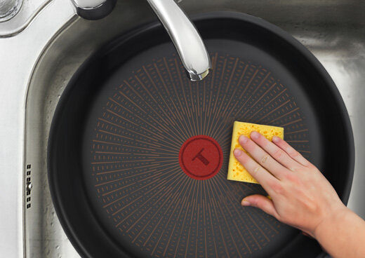 Patelnia w zlewie jest czyszczona gąbką. Na środku patelni widoczny jest czerwony okrąg z literą T.