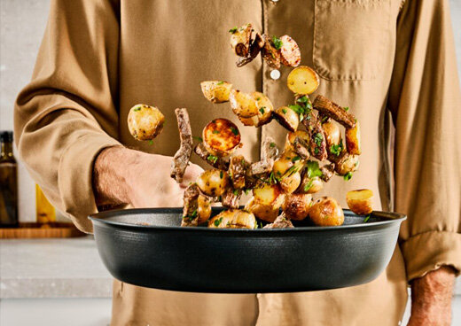 Osoba trzyma patelnię, w której unoszą się kawałki ziemniaków i mięsa. W tle widoczna jest kuchnia.