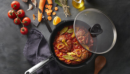Patelnia z przezroczystą pokrywką, wypełniona potrawą z mięsa i warzyw. Obok znajdują się pomidory, marchewki, przyprawy i drewniana łyżka.