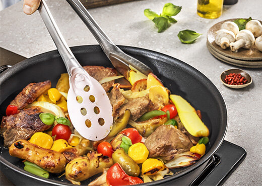 Na patelni znajdują się różnorodne warzywa i kawałki mięsa, mieszane za pomocą dwóch łopatek. W tle widoczne są przyprawy, oliwa i pieczarki.