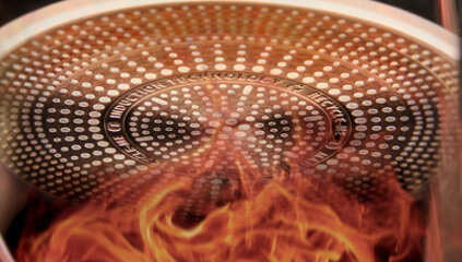 Spód patelni z widocznymi okrągłymi wzorami i napisem \'INDUCTION TECHNOLOGY\'. W tle widoczne są płomienie.