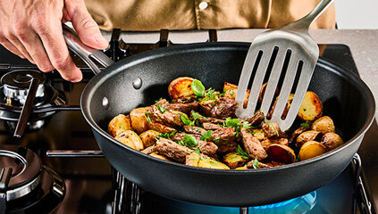 Na kuchence gazowej znajduje się patelnia z jedzeniem, w której ktoś miesza metalową łopatką. Widać kawałki mięsa i ziemniaków.