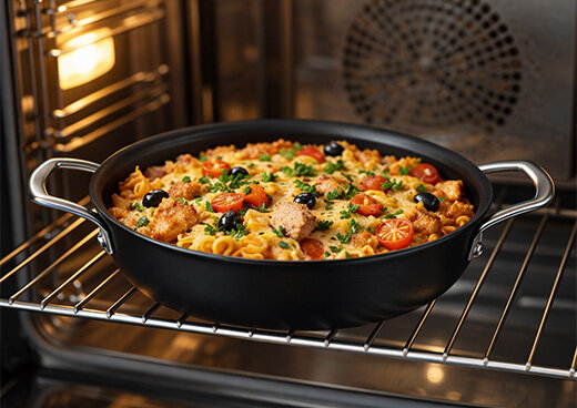 Czarna patelnia z uchwytami znajduje się w piekarniku, wypełniona zapiekanką z makaronem, pomidorami, oliwkami i ziołami.