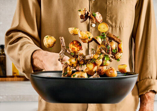 Osoba trzyma w dłoni patelnię TEFAL Excellence+ G3301902 i podrzuca jedzenie, które się na niej znajduje.