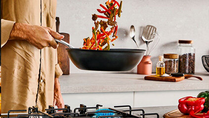 Osoba trzyma w dłoni patelnię TEFAL Excellence+ G3301902 i podrzuca jedzenie, które się na niej znajduje. W tle kuchnia.