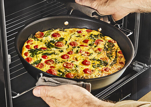 Osoba wyciąga patelnię z piekarnika, na której znajduje się danie z pomidorami i zieleniną. Patelnię trzyma za pomocą rękawic kuchennych.