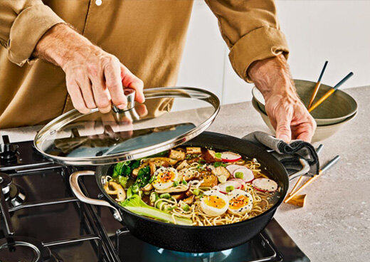 Osoba trzyma pokrywkę nad patelnią z gotującym się jedzeniem, w tym jajkami, warzywami i makaronem. Patelnię umieszczono na kuchence gazowej, obok stoi miska z pałeczkami.