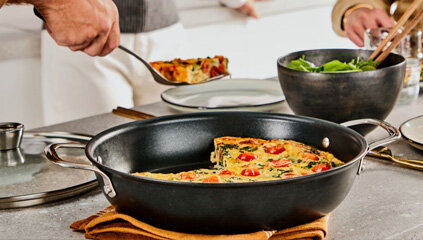 Na blacie kuchennym stoi patelnia z potrawą, obok której znajduje się miska z sałatą. Osoba nakłada kawałek potrawy na talerz.