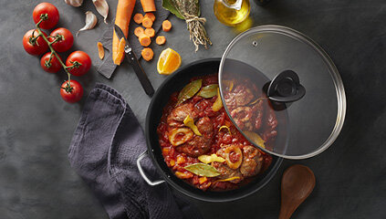 Na ciemnym tle znajduje się patelnia z przezroczystą pokrywką, w której gotuje się potrawa z mięsem i warzywami. Obok leżą pomidory, marchewki, przyprawy, oliwa oraz drewniana łyżka.