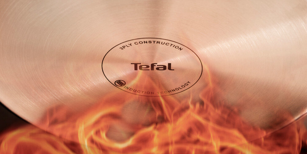 Na obrazie widoczna jest dolna część patelni z napisem \'3PLY CONSTRUCTION Tefal INDUCTION TECHNOLOGY\', otoczona płomieniami.