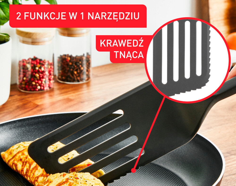 Na obrazie widoczna jest czarna łopatka kucharska z ząbkowaną krawędzią, używana do przewracania omleta na patelni. Na górze obrazu znajdują się napisy: \'2 FUNKCJE W 1 NARZĘDZIU\' oraz \'KRAWĘDŹ TNĄCA\'.