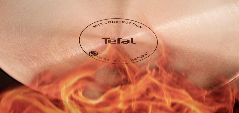 Zbliżenie na spód patelni z widocznym logo \'Tefal\' oraz napisami \'3PLY CONSTRUCTION\' i \'INDUCTION TECHNOLOGY\'. W tle widoczne są płomienie ognia.