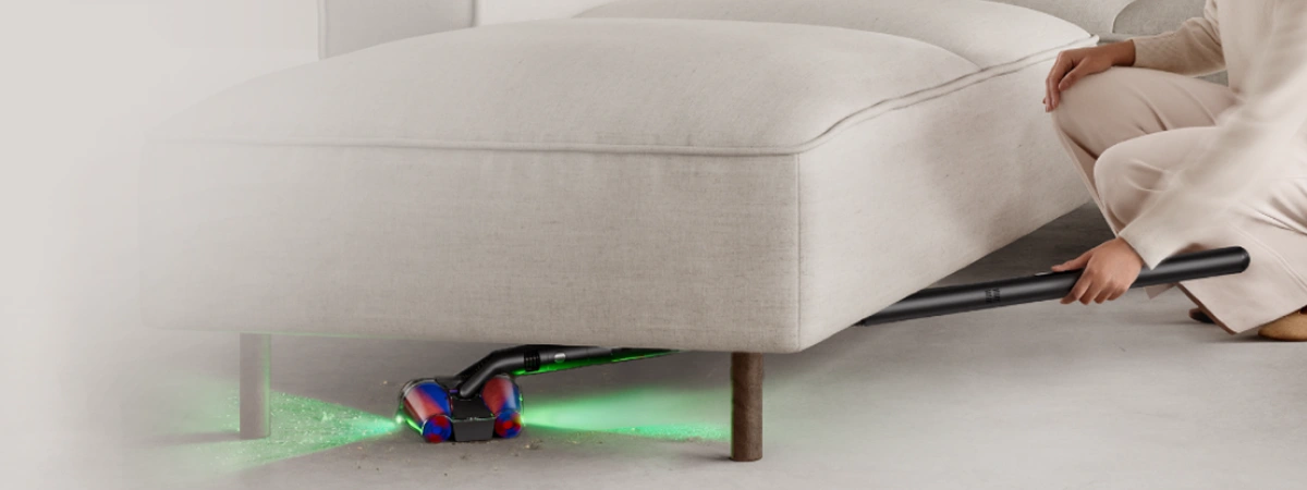 Osoba sprząta odkurzaczem trudno dostępną przestrzeń pod sofą. Odkurzacz składa się, by dotrzeć do miejsc pod meblem.
