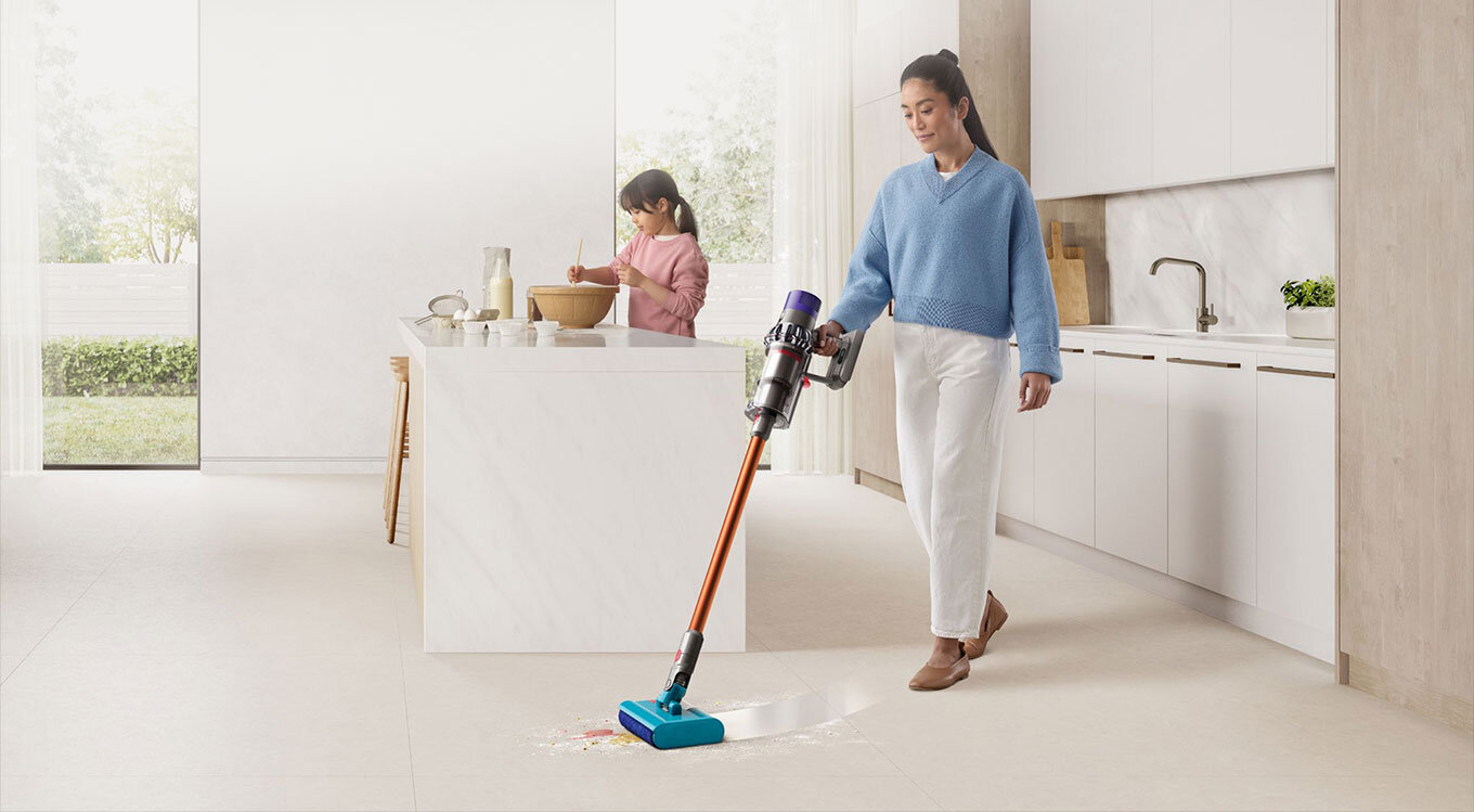 Kobieta trzyma w ręku odkurzacz Dyson V10 Submarine z końcówką do mycia podłóg. Zmywa poplamioną podłogę w jasnej kuchni.