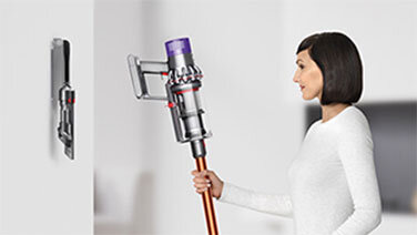Osoba trzyma odkurzacz bezprzewodowy z fioletowym elementem na górze i miedzianą rurą. W tle widoczny jest uchwyt ścienny na odkurzacz.