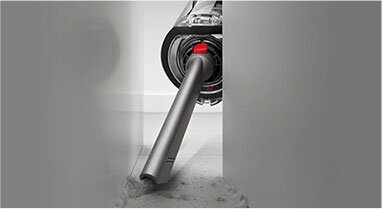 Odkurzacz z długą, wąską końcówką ssącą, czyszczący przestrzeń między dwoma powierzchniami. Widoczny jest kurz na podłodze, a urządzenie ma elementy w kolorze srebrnym i czerwonym.