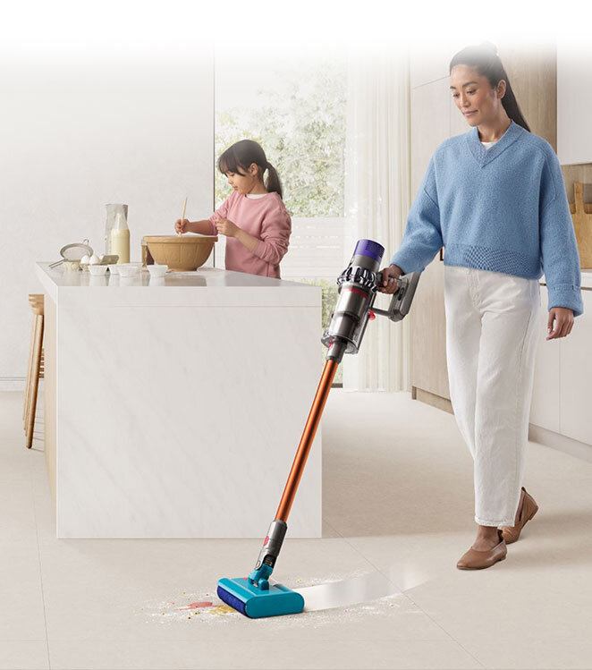 Osoba używa odkurzacza bezprzewodowego z pomarańczową rurą i niebieską końcówką na jasnej podłodze. W tle widać kuchnię z osobą mieszającą w misce na blacie.