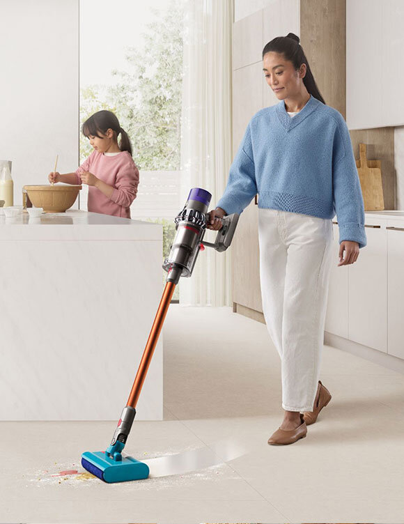 Osoba używa odkurzacza bezprzewodowego z pomarańczową rurą i niebieską końcówką na jasnej podłodze. W tle widać kuchnię i osobę mieszającą coś w misce.