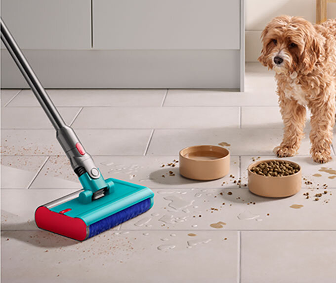 Na podłodze widać odkurzacz z turkusową głowicą i niebieską szczotką, który sprząta rozsypane jedzenie i rozlaną wodę. Obok stoją dwie miski, a w tle znajduje się pies.