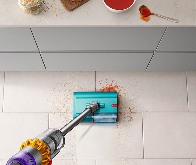 Odkurzacz z turkusową końcówką sprząta rozlany sos na jasnej podłodze. Na blacie widać miskę z płatkami i rozlany sos obok łyżki.