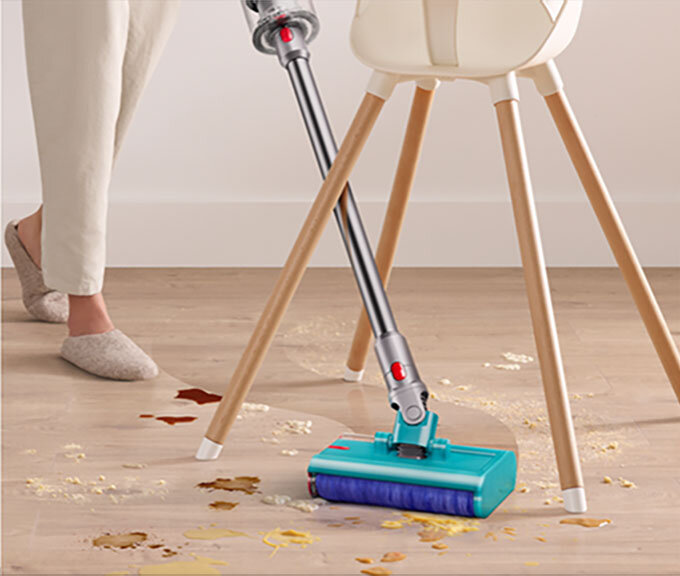 Osoba używa odkurzacza z turkusową głowicą do czyszczenia podłogi z rozsypanych płatków i płynów. W tle widoczne są nogi krzesła i fragment podłogi.