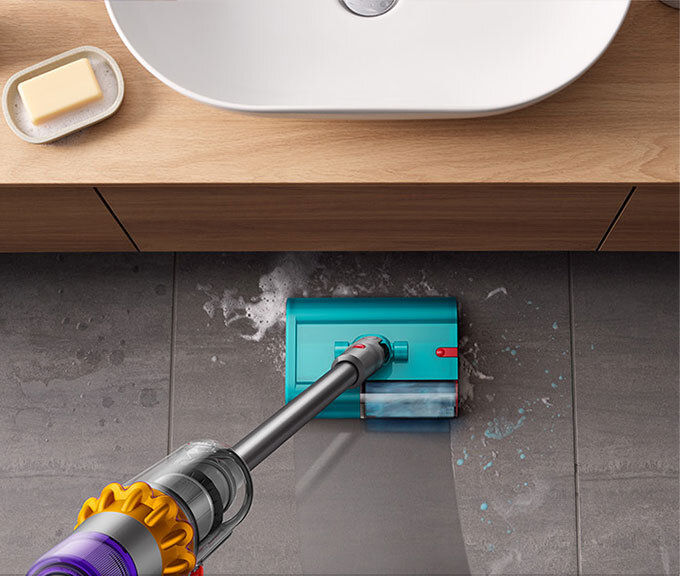 Na obrazie widać odkurzacz myjący podłogę z szarych płytek, z widoczną wodą i pianą. W tle znajduje się umywalka i mydelniczka z kostką mydła.