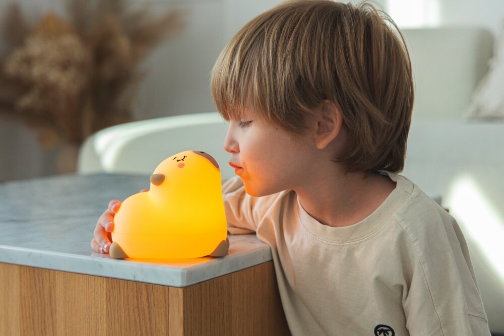 Lampka nocna INNOGIO Giococo the Capybara GIO-157 Mały chłopiec przybliża twarz do świecącej lampki nocnej w kształcie kapibary, która stoi na drewnianej szafce z marmurowym blatem.