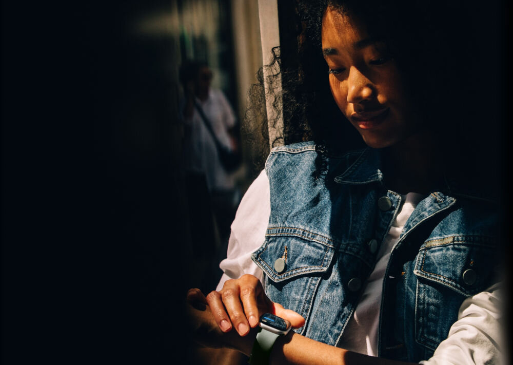 Apple Watch 11 GPS 42mm koperta z aluminium (różowe złoto) + pasek sportowy rozmiar S/M (łagodny róż) kobieta ma założony zegarek na ręce drugą dłonią dotyka go czarne tło i widoczne odbicie drugiej osoby sieć komórkowa wygoda gesty znajdowanie iPhone sugestie