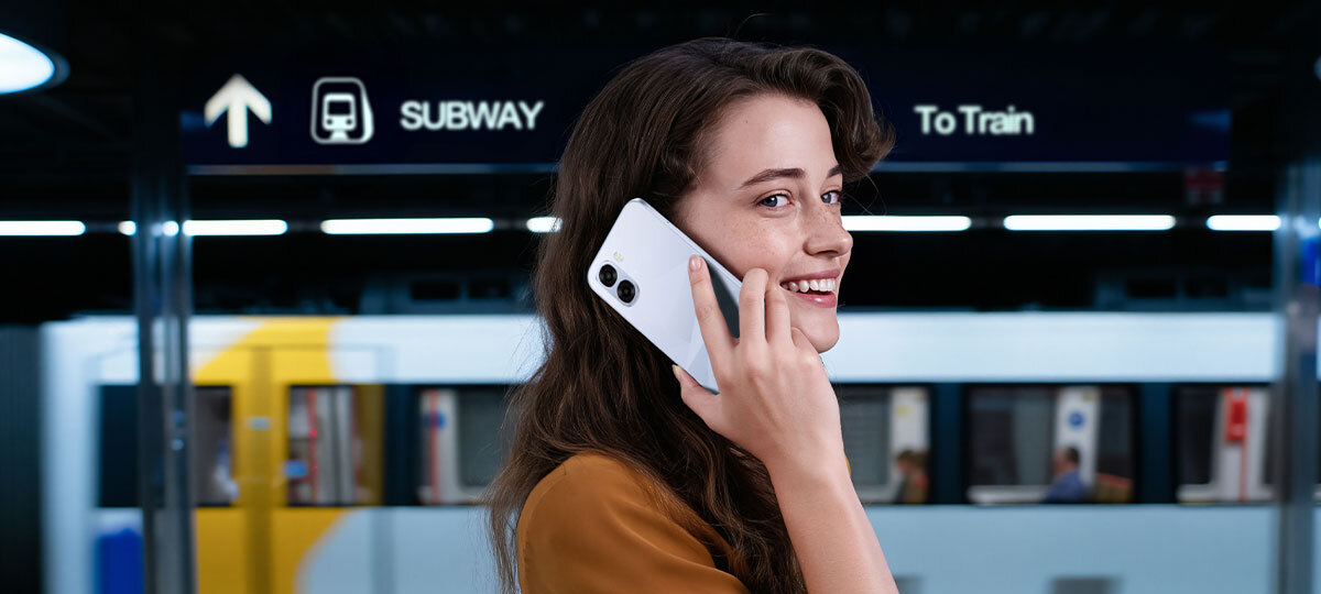 Osoba trzymająca smartfon przy uchu stoi na tle stacji metra. W tle widoczne są znaki z napisami \'SUBWAY\' i \'To Train\'.