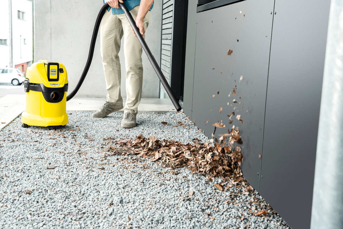 Osoba używa żółtego odkurzacza warsztatowego do usuwania liści z powierzchni pokrytej żwirem. Na odkurzaczu widoczne jest logo \'KARCHER\'.