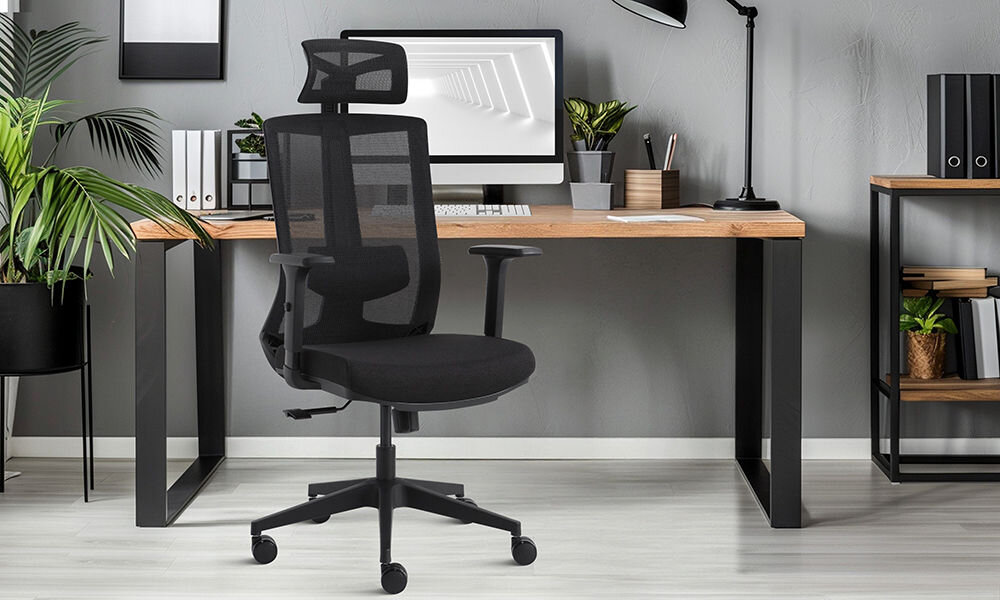 Czarny fotel biurowy z siatkowym oparciem i zagłówkiem stoi przed drewnianym biurkiem z metalowymi nogami. Na biurku znajdują się monitor, klawiatura, doniczki z roślinami i segregatory.