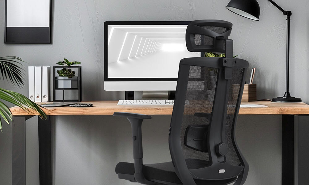 Czarny fotel biurowy z siatkowym oparciem stoi przed biurkiem z komputerem i lampką. Na biurku znajdują się również książki i roślina w doniczce.