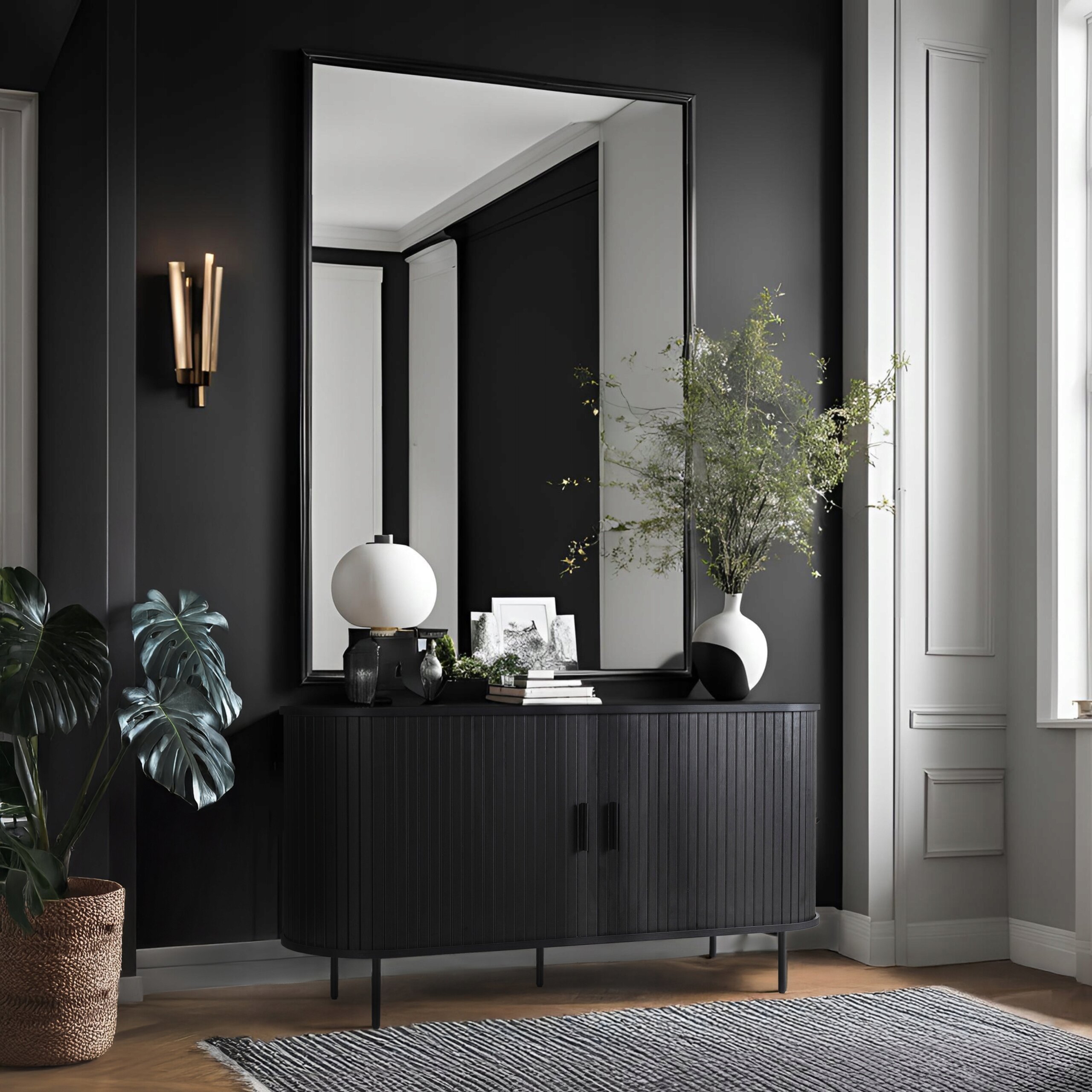 Czarna komoda z lamelami stoi w eleganckim wnętrzu, na niej znajduje się lampa, książki i wazon z zieloną rośliną. W tle duże lustro i ściana w ciemnym kolorze z dekoracyjnym kinkietem.