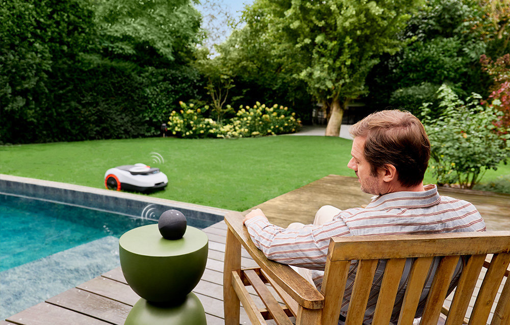 Robot koszący SEGWAY NAVIMOW i220E ogród trawnik robot kosi trawę na pierwszym planie osoba siedzi na krześle ogrodowym w tle drzewa krzewy współpracuje z Amazon Alexa i Google Home sterowanie robotem za pomocą prostych komend głosowych