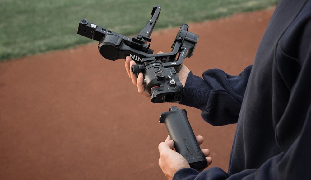Gimbal DJI RS 5 Ręce operatora montującego lub demontującego uchwyt gimbalów na boisku sportowym, trzymające oddzielony element w formie cylindra