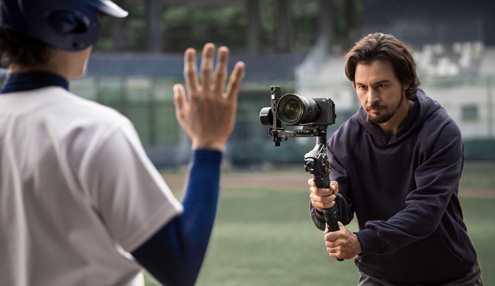 Gimbal DJI RS 5 Operator filmujący zawodnika baseballu na boisku, trzymający gimbal z zamontowanym aparatem na rozłożonym statywie