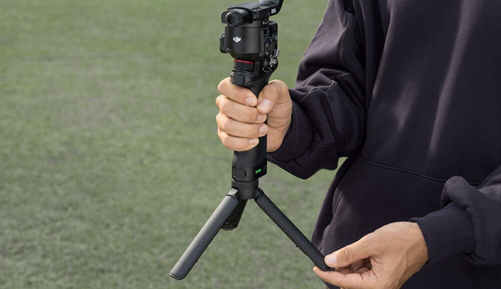 Gimbal DJI RS 5 Combo Osoba w bluzie trzymająca gimbal z zamontowanym aparatem, chwytając jednocześnie rozkładane nogi statywu