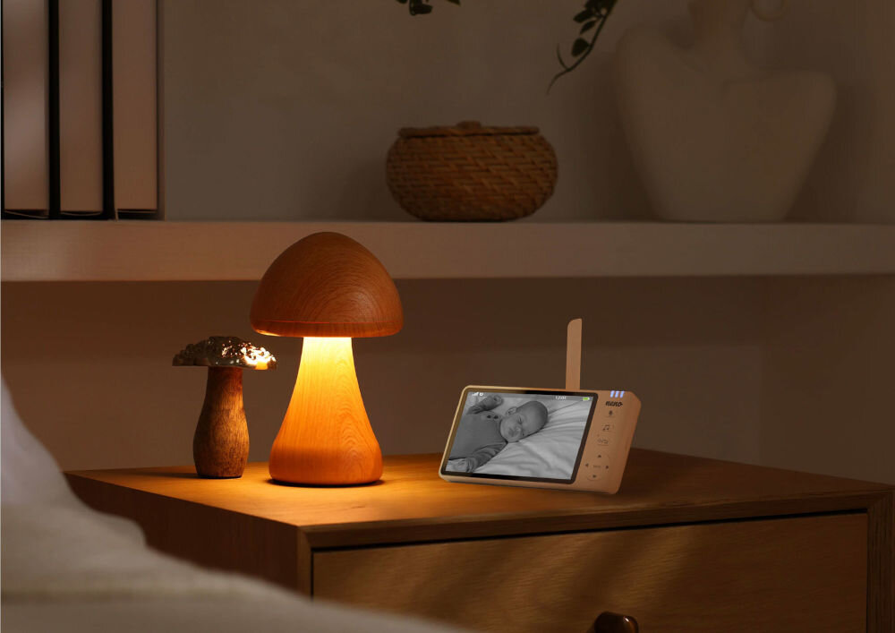 Na drewnianej szafce nocnej stoi lampka w kształcie grzyba oraz elektroniczna niania z wyświetlaczem pokazującym śpiące dziecko. W tle widoczny jest koszyk na półce.