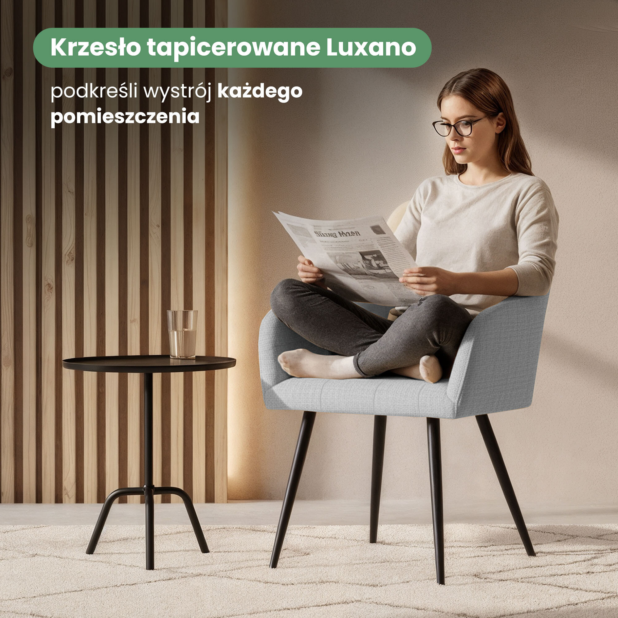 Osoba siedzi na jasnoszarym, tapicerowanym krześle z czarnymi nogami, trzymając gazetę. Obok znajduje się mały, czarny stolik z przezroczystą szklanką, a w tle widoczna jest drewniana ściana z pionowymi panelami.