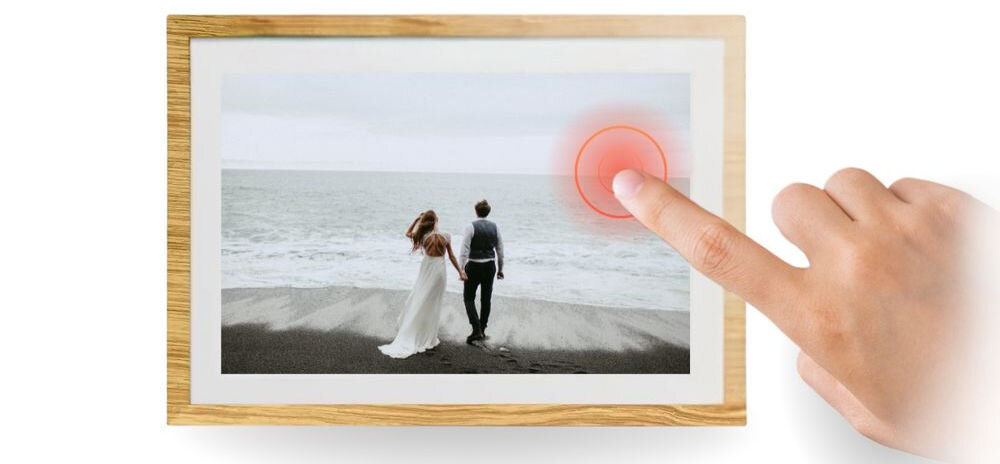 Na obrazie widoczna jest cyfrowa ramka z wyświetlonym zdjęciem pary spacerującej po plaży. Obok ramki znajduje się dłoń wskazująca na ekran, z zaznaczonym czerwonym okręgiem dotykowym.