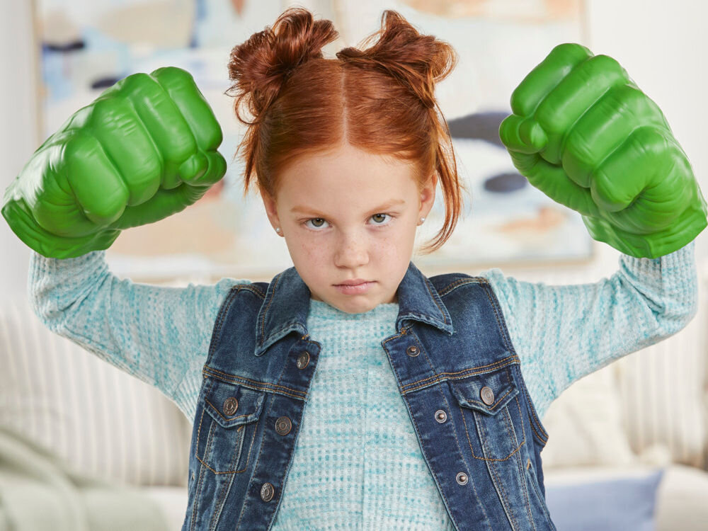 Rękawice HASBRO Marvel Avengers Gamma Smash Pięści Hulka F93325L0 Zbliżenie na zieloną rękawicę Hulka trzymaną przez dziecko w geście uderzenia. Detale zabawki podkreślają fakturę skóry i zaciśniętą pięść. W tle widoczne błękitne niebo z białymi grafikami w stylu komiksowym.