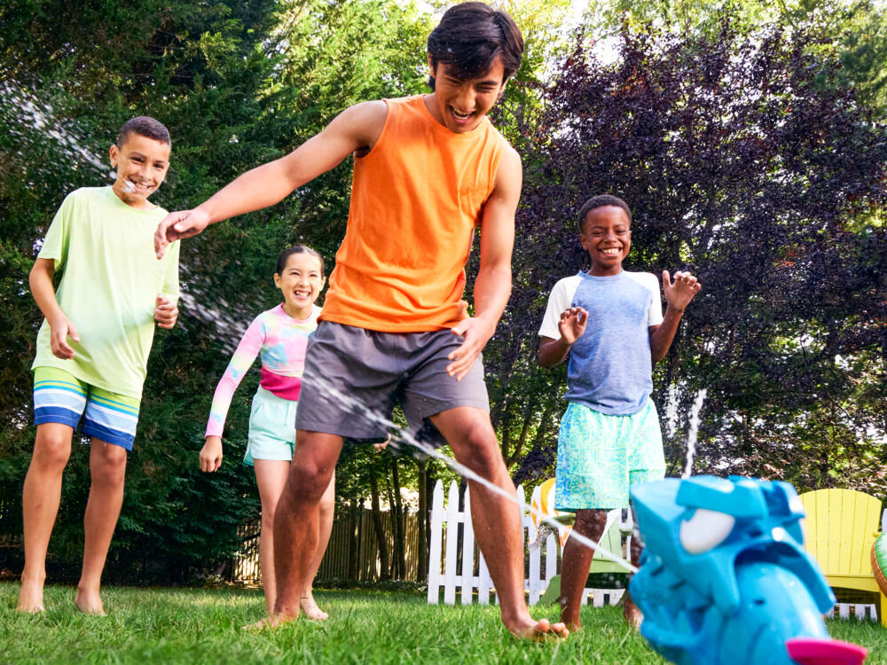 Wyrzutnia NERF Super Soaker Hop & Chomp G21485L0 Grupa uśmiechniętych dzieci bawiących się w ogrodzie, przeskakujących nad strumieniem wody tryskającym z zraszacza w kształcie rekina.