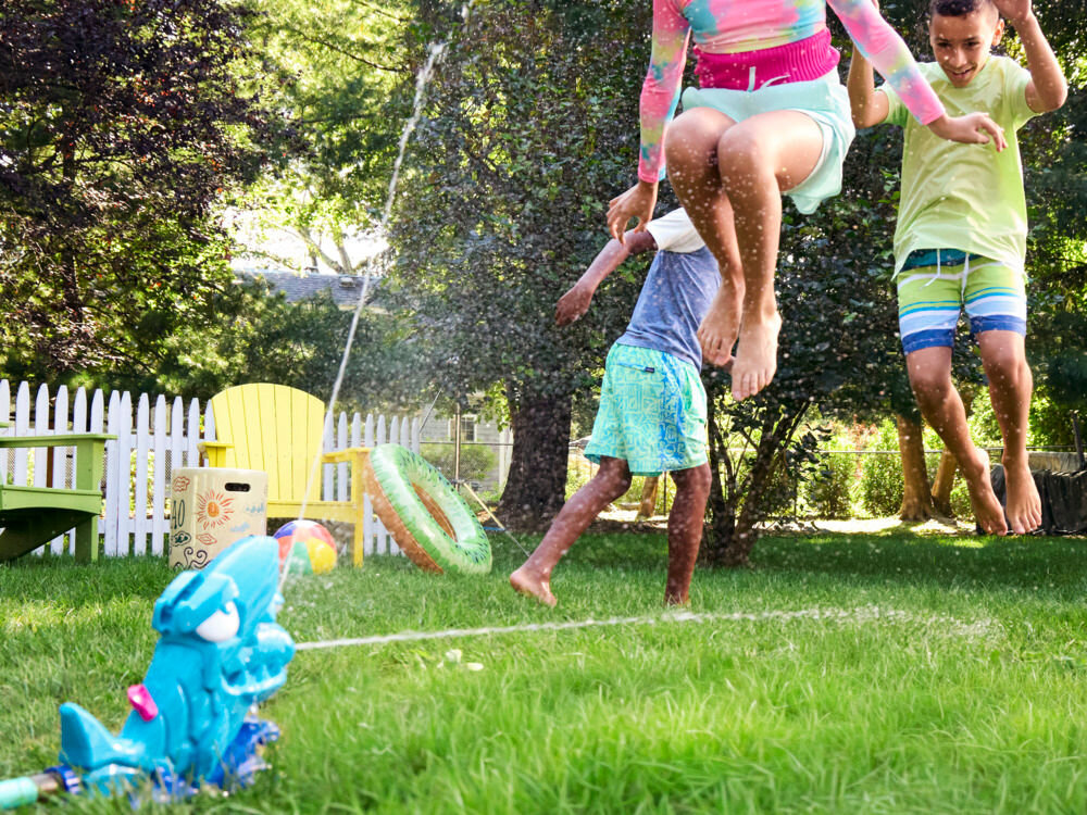 Wyrzutnia NERF Super Soaker Hop & Chomp G21485L0 Dzieci w strojach kąpielowych skaczą nad strumieniem wody na trawniku podczas zabawy zraszaczem Nerf Super Soaker.