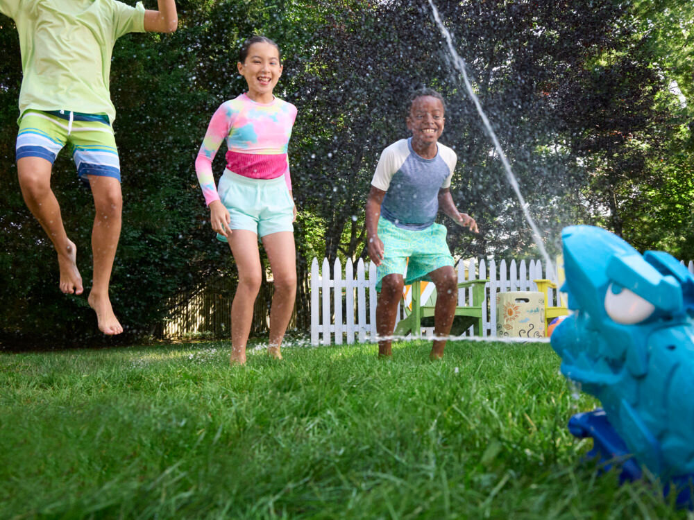 Wyrzutnia NERF Super Soaker Hop & Chomp G21485L0 Radosne dzieci przeskakujące przez wodę tryskającą z obrotowego zraszacza w kształcie rekina podczas letniej zabawy na dworze.