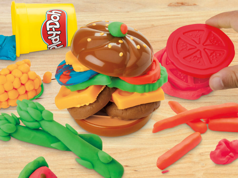 Na drewnianym stole znajduje się zestaw ciastoliny w kształcie hamburgera z dodatkami, obok leży żółty pojemnik z napisem \'Play-Doh\'. Widać również różnokolorowe elementy przypominające warzywa i dodatki do potraw.