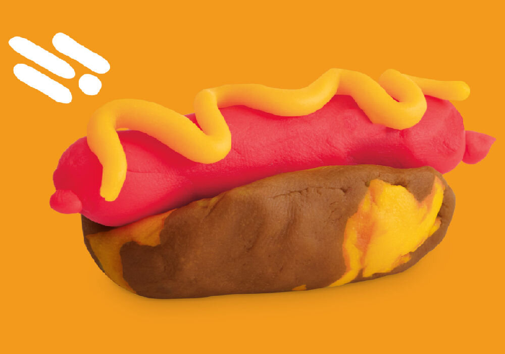 Na pomarańczowym tle znajduje się model hot doga wykonany z ciastoliny, z brązową bułką, różową parówką i żółtym sosem. W lewym górnym rogu widoczne są trzy białe kreski.