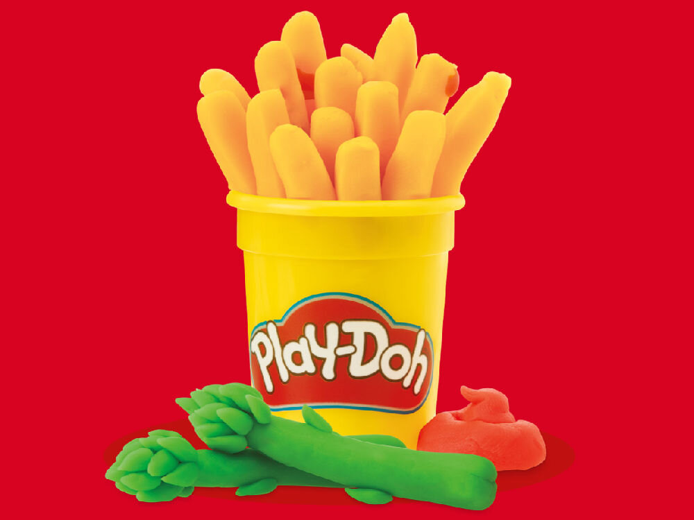 Na czerwonym tle znajduje się żółte pudełko z logo \'Play-Doh\', wypełnione pomarańczowymi kształtami przypominającymi frytki. Obok pudełka leżą zielone kształty przypominające szparagi oraz mała czerwona kulka.