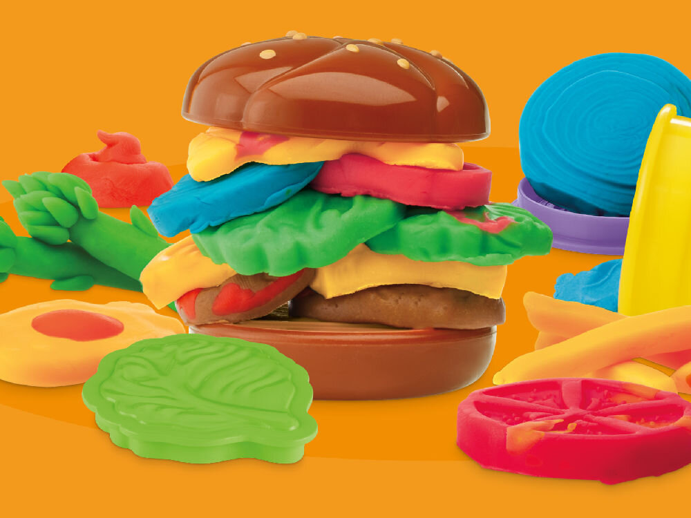 Na obrazie widoczny jest kolorowy hamburger z ciastoliny, złożony z różnych warstw w różnych kolorach, takich jak brązowy, zielony, żółty, czerwony i niebieski. W tle znajdują się inne elementy z ciastoliny, przypominające warzywa i dodatki.